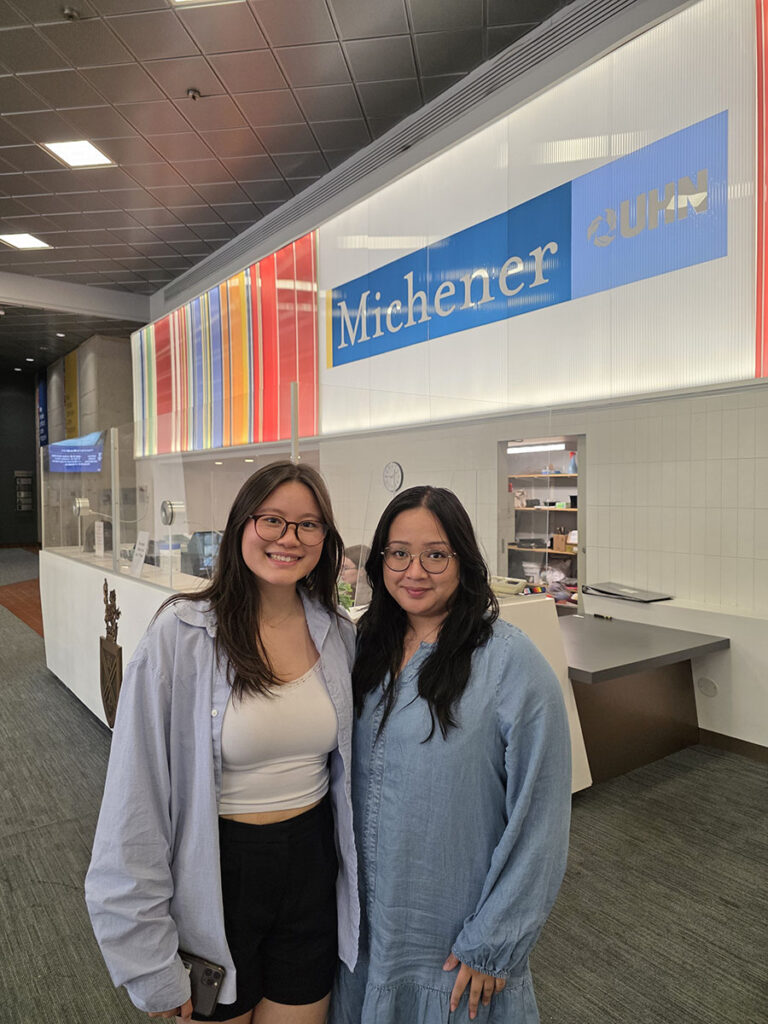 Two women stand smiling in front of the Michener sign inside a modern building lobby with colorful wall decor.